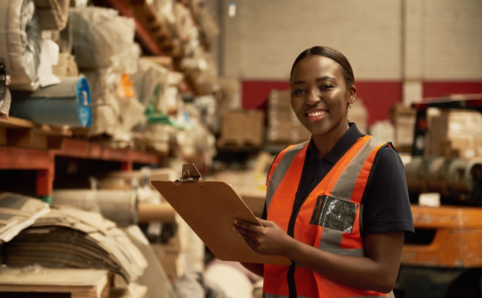Woman wearing an orange safety vest holds a clipboard in a warehouse.