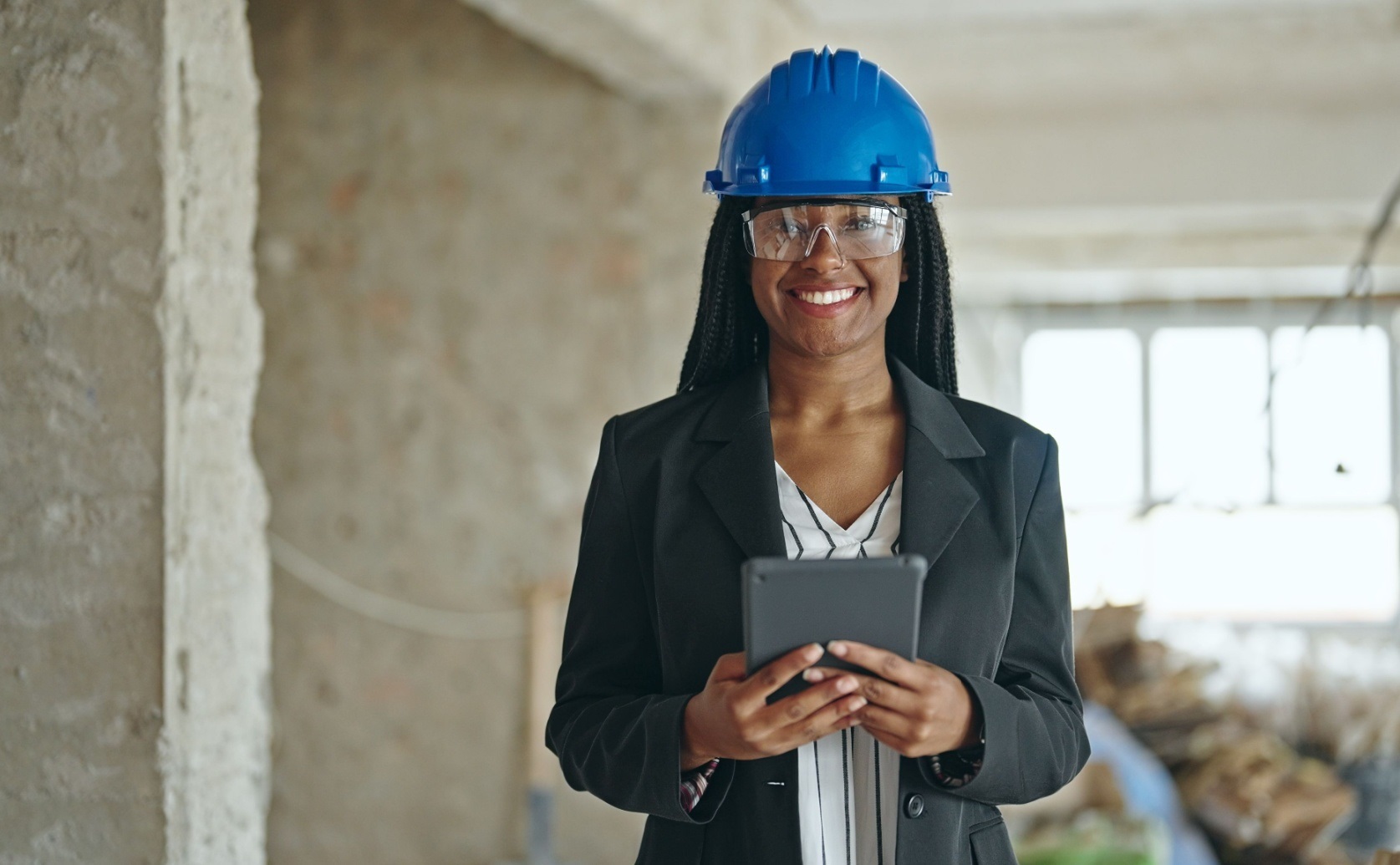 Woman wearing a blue hard hat and safety glasses holds a tablet inside an unfinished building.