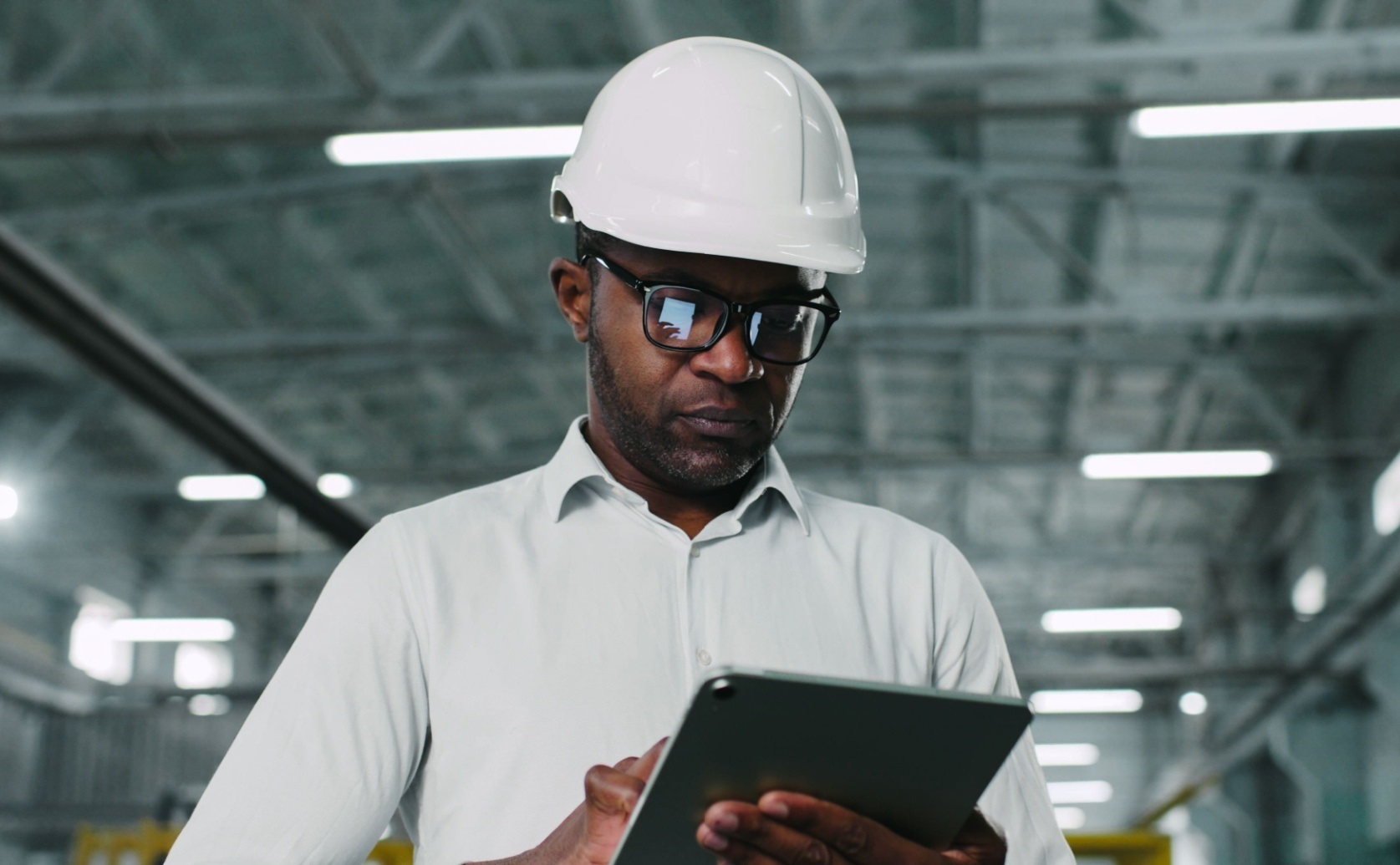 Man wearing a hard hat and safety glasses uses a tablet in a factory.