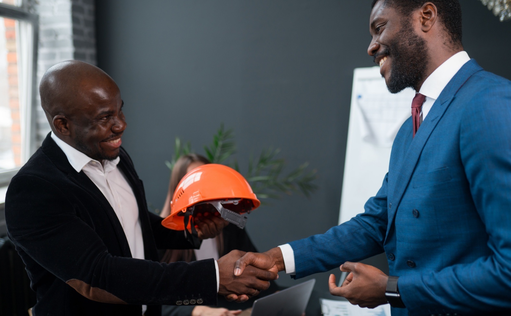 Two men shake hands while one holds an orange hard hat.