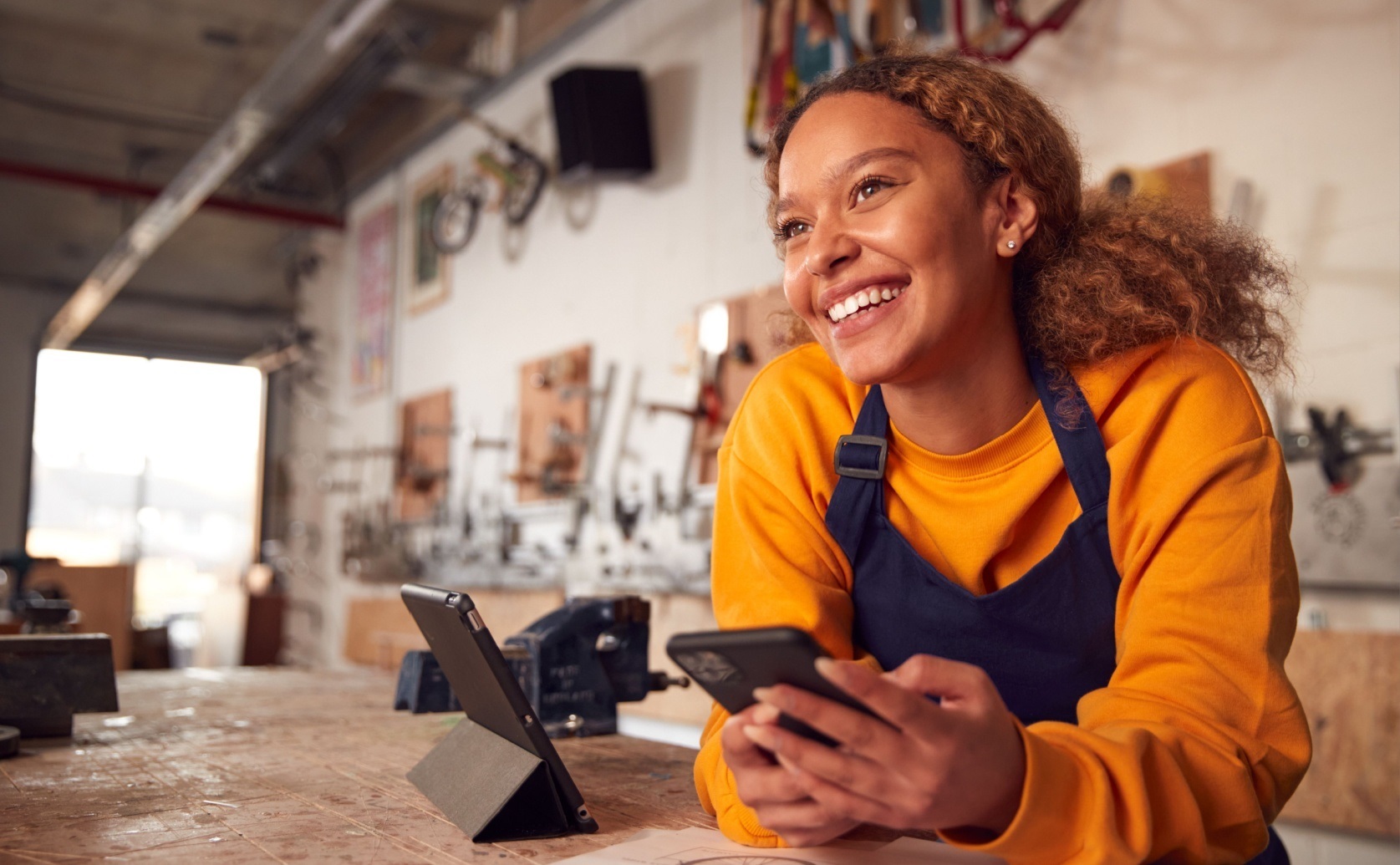 Woman wearing an apron holds a phone at a workshop counter.
