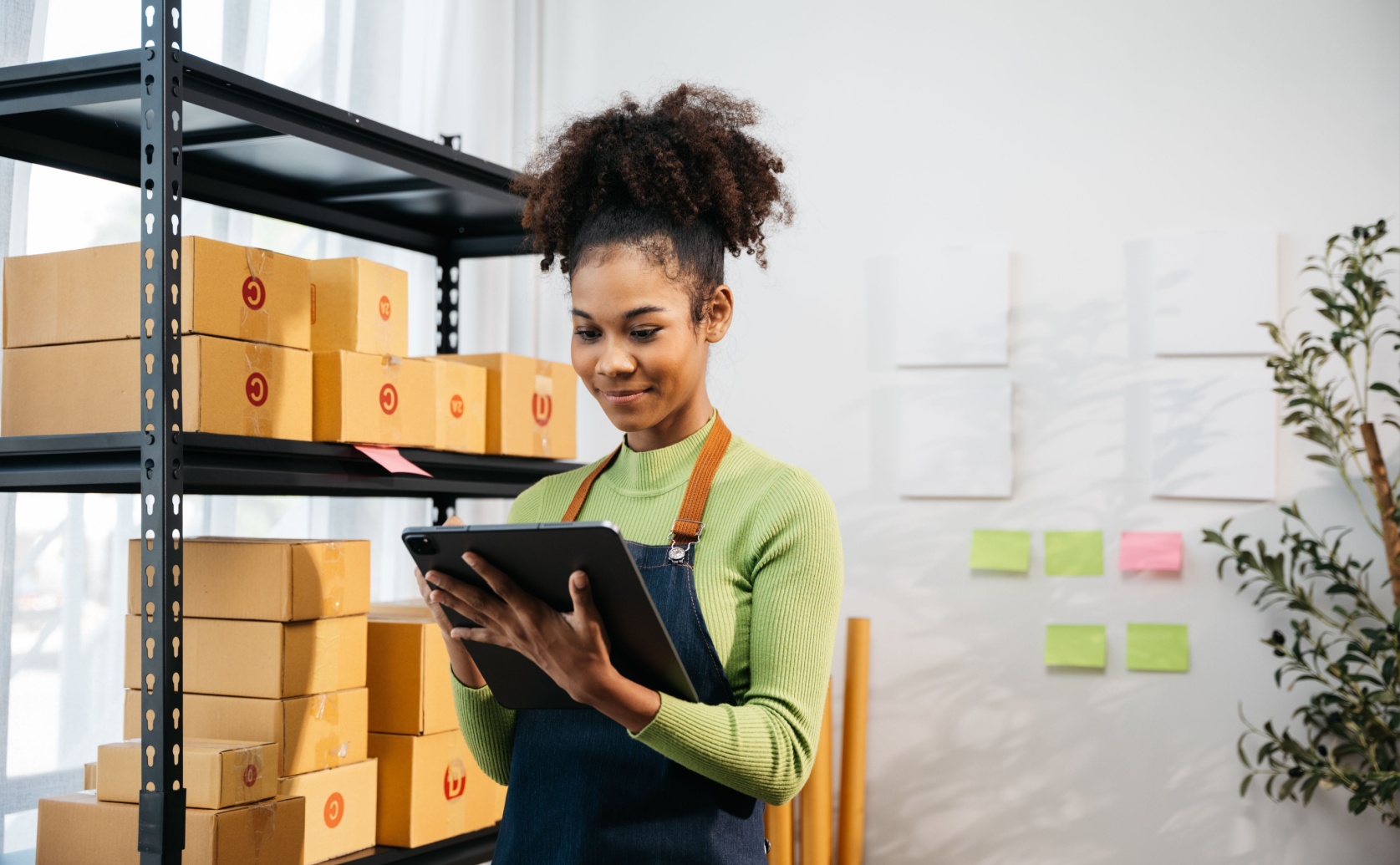 Woman wearing an apron uses a tablet beside shelves of parcels.