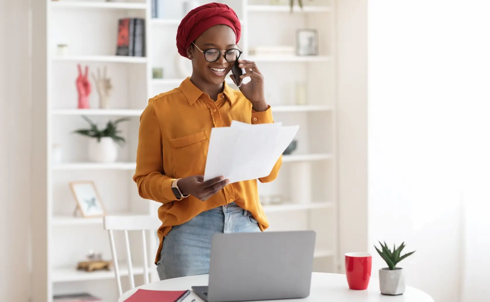 Woman holds paperwork and talks on a phone beside an open laptop.