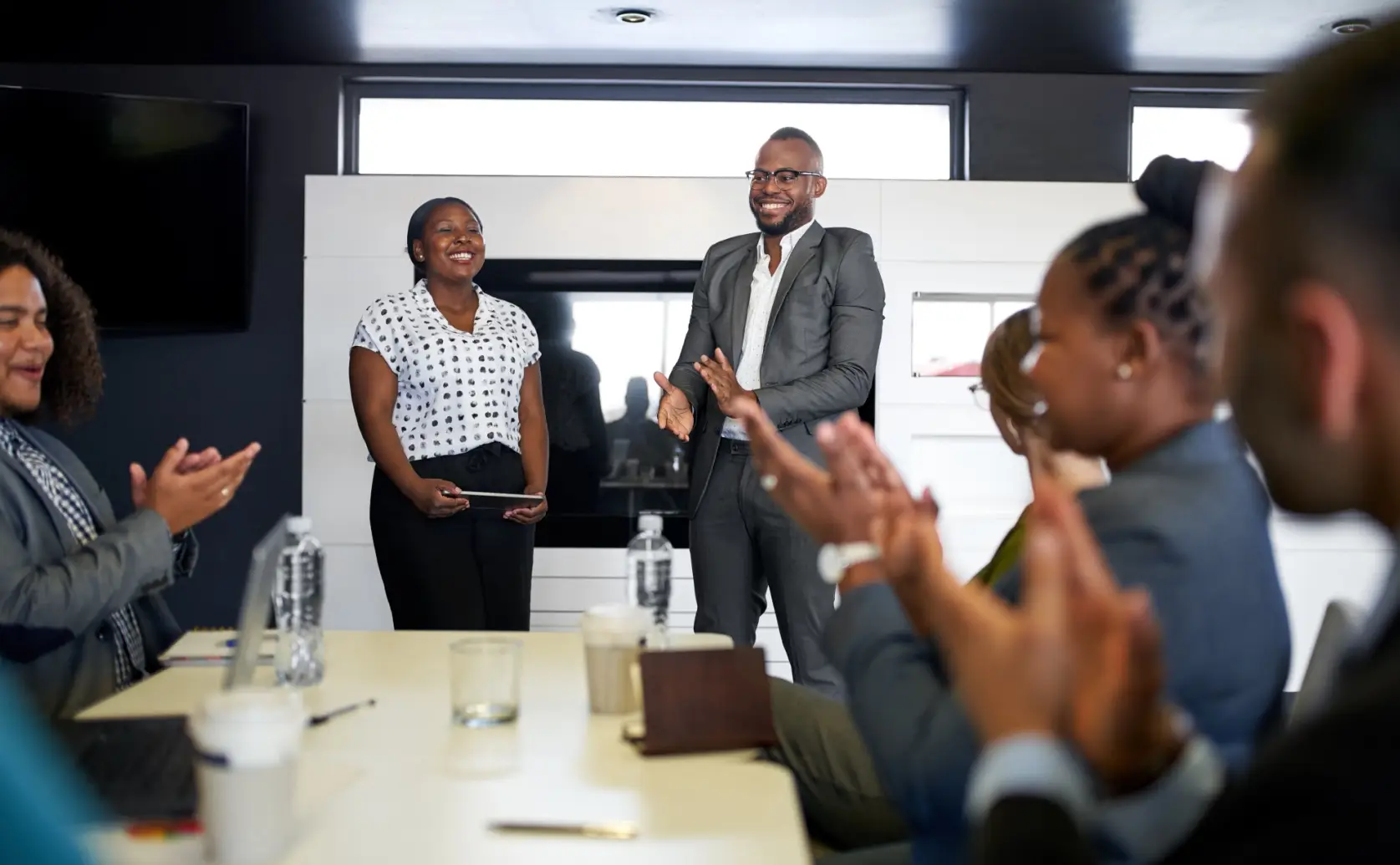 Colleagues applaud two people standing at the end of a conference table.