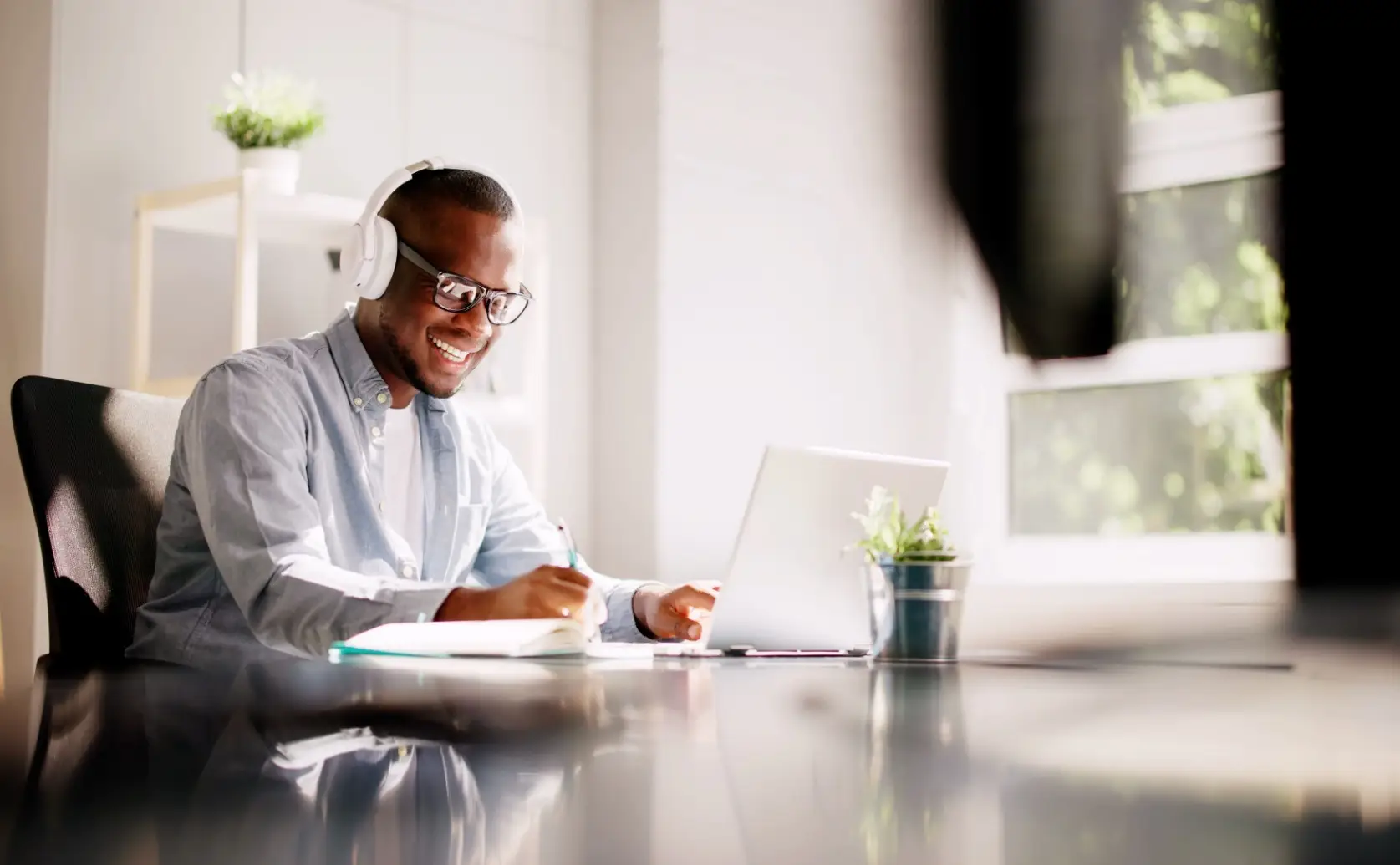 Man works on a laptop and writes in a notebook at a table.