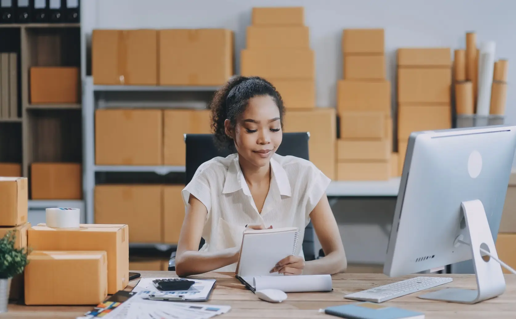 Woman reviews documents at a computer with cardboard boxes behind her.