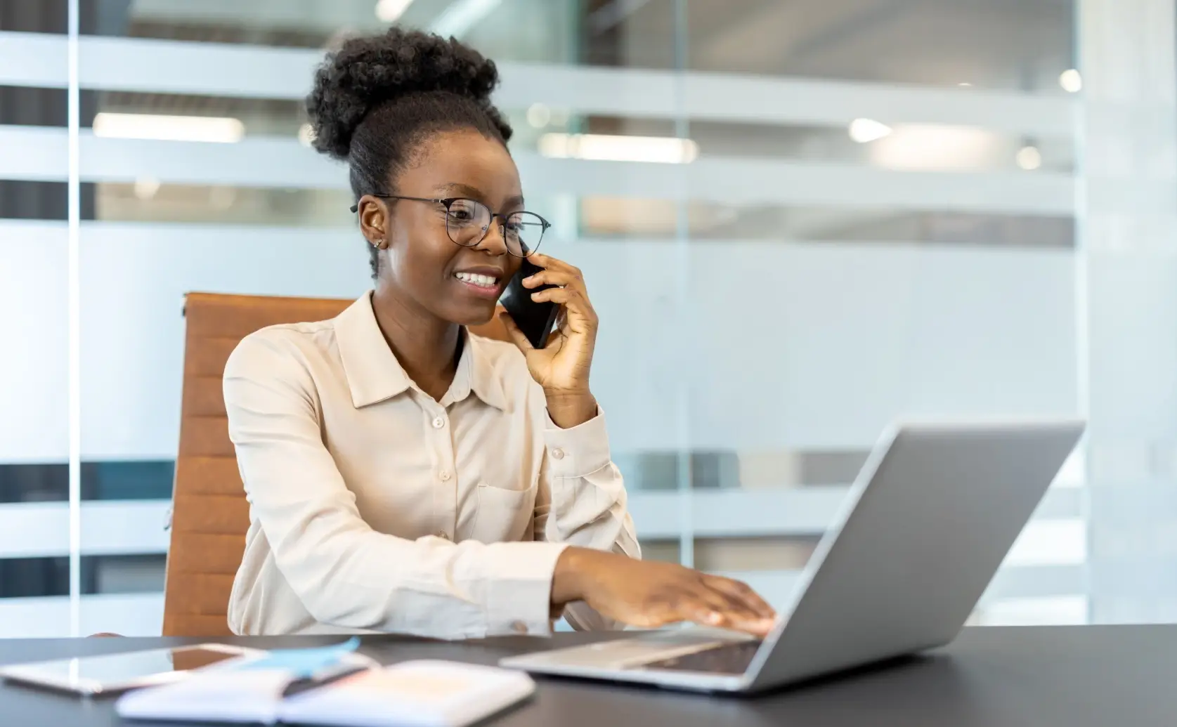 Woman talks on a phone while using a laptop at an office desk.