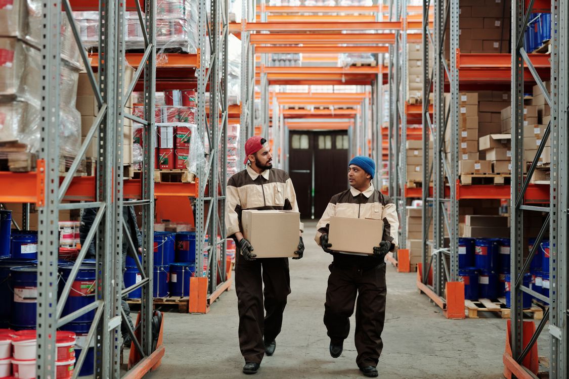 Two warehouse workers carrying boxes along a storage aisle.